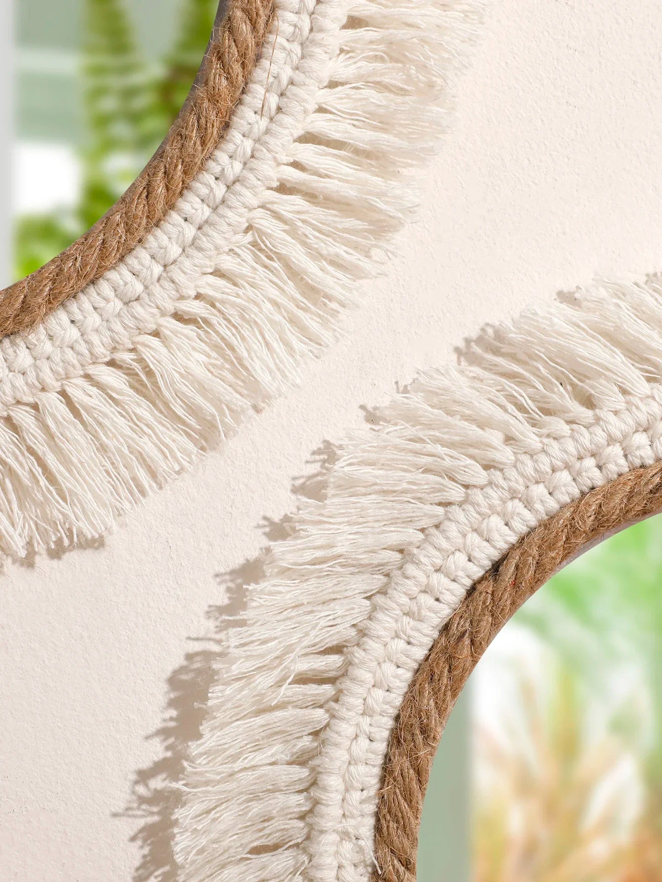 Close-up of a round mirror with macrame frame and fringes, blurred greenery in the background
