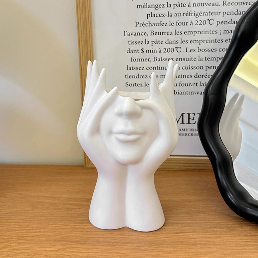 White ceramic sculpture of a face with raised hands on a wooden surface, with a mirror reflecting the sculpture.