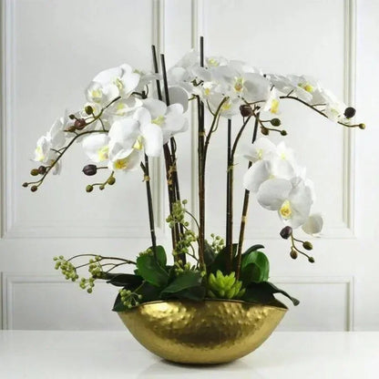 Decorative arrangement of white flowers and green leaves in a gold bowl against a white paneled wall.