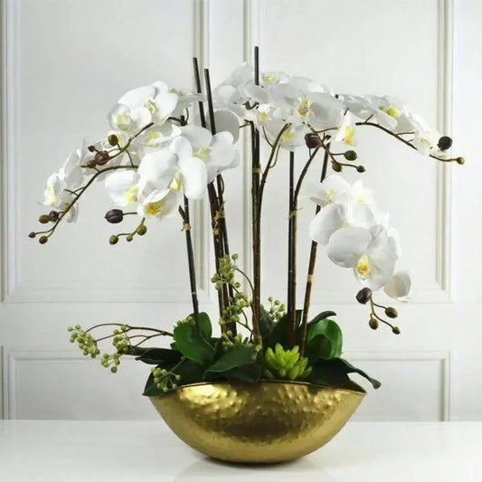 Decorative arrangement of white flowers and green leaves in a gold bowl against a white paneled wall.