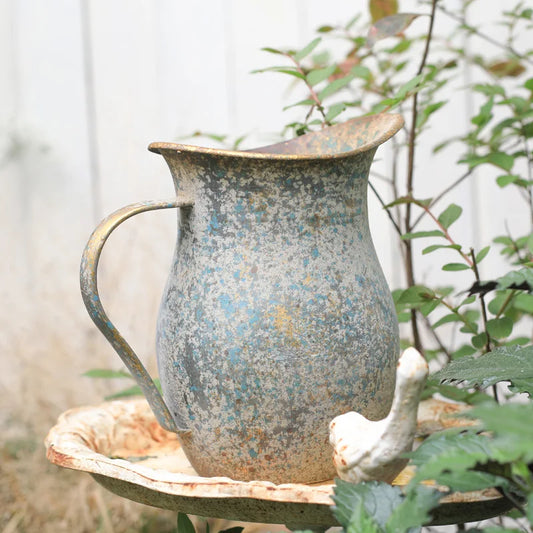 Rustic metal pitcher on a wooden surface with greenery in the background